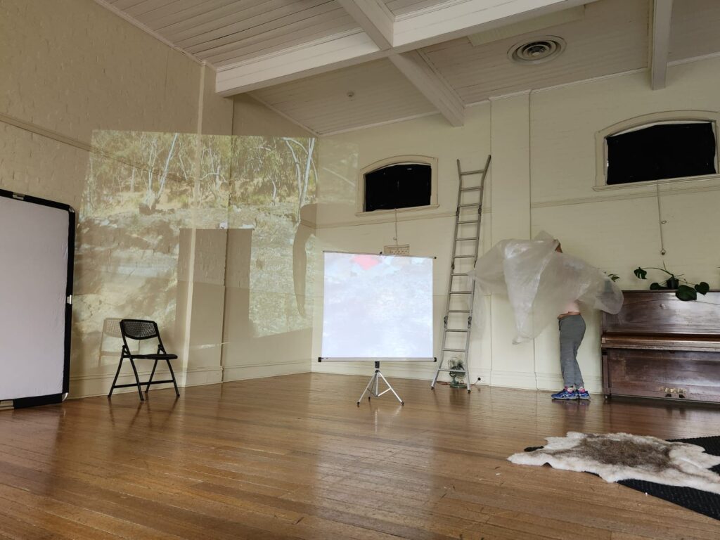 A studio space with projections of nature, a screen, a ladder and a person standing at the back wall with a bubble wrap head piece.