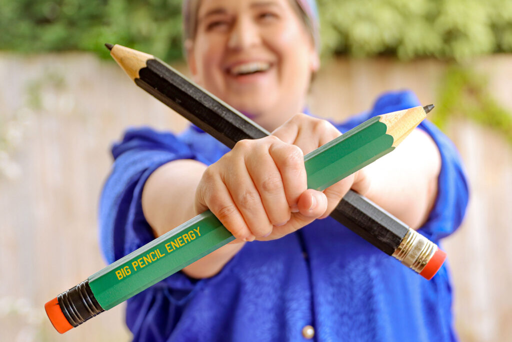 Megan holds up two giant pencils crossing each other. The pencil says BIG PENCIL ENERGY. Her smiling face is blurred in the background.